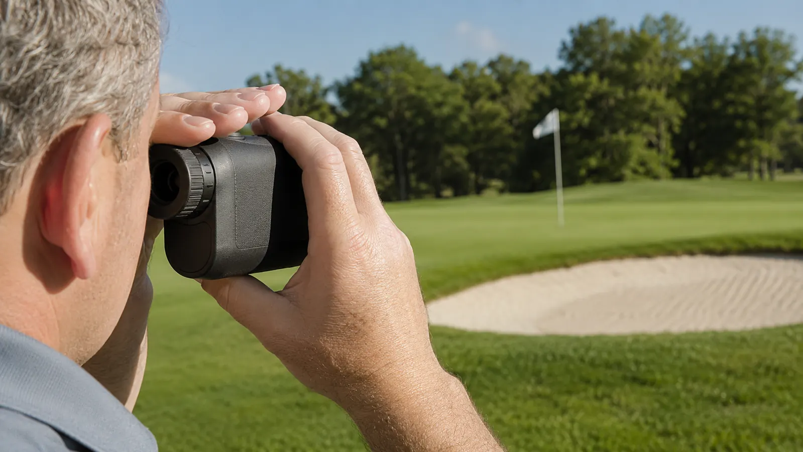 A golfer raising a rangefinder to his eye to measure a yardage to the flag on a sunny day