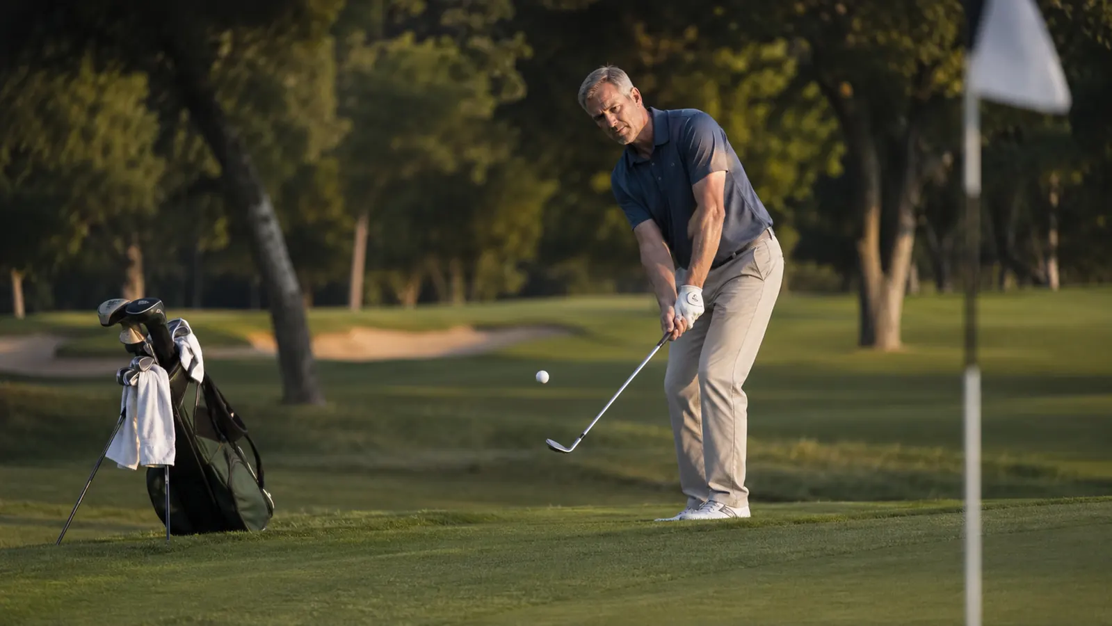 A golfer in a polo shirt lining up a pitch shot from the short grass right of a green, club held up to check the line