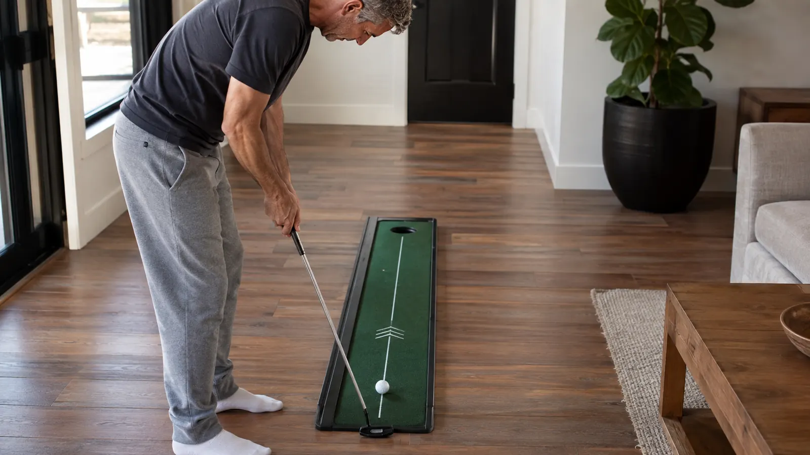 A person's hand gripping a putter as they roll a golf ball down the center line of the SKLZ putting practice strip at home